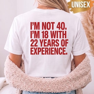 以下が含まれることがあります： 背面に赤い文字で「I'M NOT 40. I'M 18 WITH 22 YEARS OF EXPERIENCE.」とプリントされた白い半袖Tシャツ。右上の金色のグラフィックには、白い大文字で「UNISEX」と書かれています。