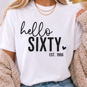Puede incluir: Una camiseta blanca de manga corta con el texto negro "hello SIXTY.", un pequeño icono de corazón y "EST. 1966" impreso debajo. Un gráfico dorado en forma de pincelada en la esquina superior derecha contiene el texto blanco "UNISEX".