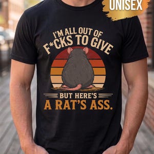 Może przedstawiać: Czarny t-shirt z krótkim rękawem z grafiką szczura widzianego od tyłu na tle zachodzącego słońca. Napis głosi I'M ALL OUT OF F*CKS TO GIVE BUT HERE'S A RAT'S ASS. Złota grafika w kształcie pociągnięcia pędzla w rogu zawiera słowo UNISEX.
