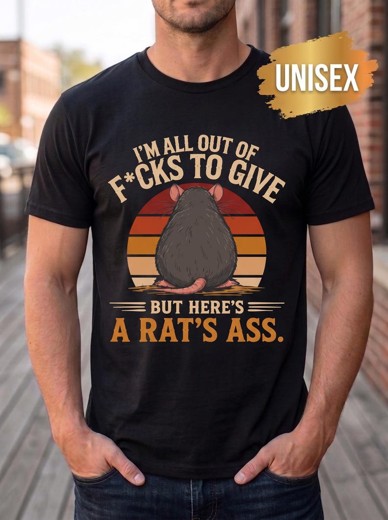 May include: A black short-sleeved t-shirt featuring a graphic of a rat viewed from behind against a sunset background. The text reads I'M ALL OUT OF F*CKS TO GIVE BUT HERE'S A RAT'S ASS. A gold-colored brushstroke graphic in the corner says UNISEX.