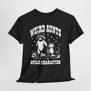 May include: Black t-shirt with a graphic featuring a raccoon and opossum, with the text "WEIRD AUNTS BUILD CHARACTER" in white. The design includes stars and grass details, creating a whimsical and fun aesthetic.