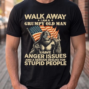 Op de afbeelding: Een zwart T-shirt met korte mouwen en een afbeelding van een harig wezen met een zonnebril in de kleuren van de Amerikaanse vlag, dat een Amerikaanse vlag vasthoudt en een handgebaar maakt. De tekst luidt: WALK AWAY I AM A GRUMPY OLD MAN I HAVE ANGER ISSUES AND A SERIOUS DESLIKE FOR STUPID PEOPLE.