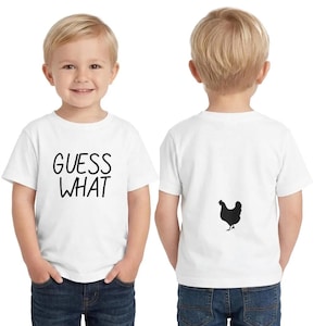 May include: White toddler t-shirt with the text "GUESS WHAT" in black on the front. The back of the shirt features a black silhouette of a chicken. The child is wearing blue jeans.
