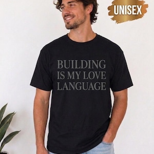 May include: A black short-sleeved t-shirt featuring the text "BUILDING IS MY LOVE LANGUAGE" printed in a grey serif font across the chest. The shirt has a crew neckline and a standard fit, with a gold brushstroke graphic in the corner.