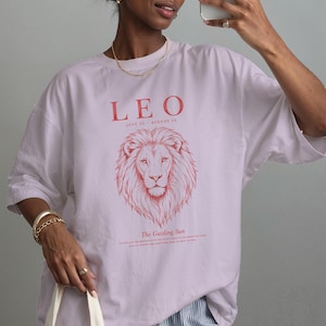 K&ouml;nnte beinhalten: Ein hellviolettes T-Shirt mit einer roten Grafik eines L&ouml;wenkopfes. Der Text lautet LEO, JULY 23 - AUGUST 22 und The Guiding Sun. Das T-Shirt hat einen Rundhalsausschnitt, kurze &Auml;rmel, eine lockere Passform und ein aufgedrucktes Design auf der Vorderseite.