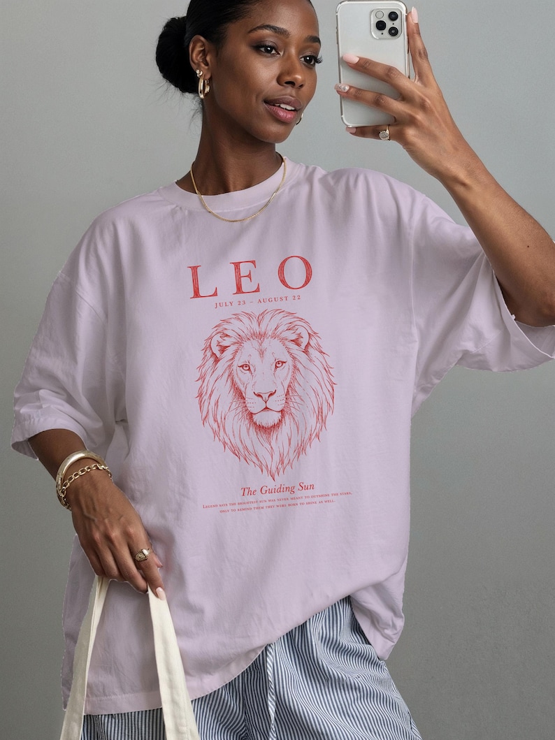 K&ouml;nnte beinhalten: Ein hellviolettes T-Shirt mit einer roten Grafik eines L&ouml;wenkopfes. Der Text lautet LEO, JULY 23 - AUGUST 22 und The Guiding Sun. Das T-Shirt hat einen Rundhalsausschnitt, kurze &Auml;rmel, eine lockere Passform und ein aufgedrucktes Design auf der Vorderseite.