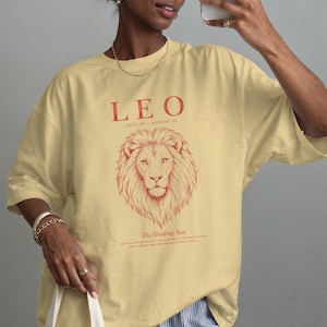 K&ouml;nnte beinhalten: Ein hellgelbes Kurzarm-T-Shirt mit einer roten Grafik eines L&ouml;wenkopfes. Der Text lautet LEO, JULY 23 - AUGUST 22 und The Guiding Sun, mit kleinerem Text darunter. Das T-Shirt hat einen Rundhalsausschnitt und eine lockere, entspannte Passform.