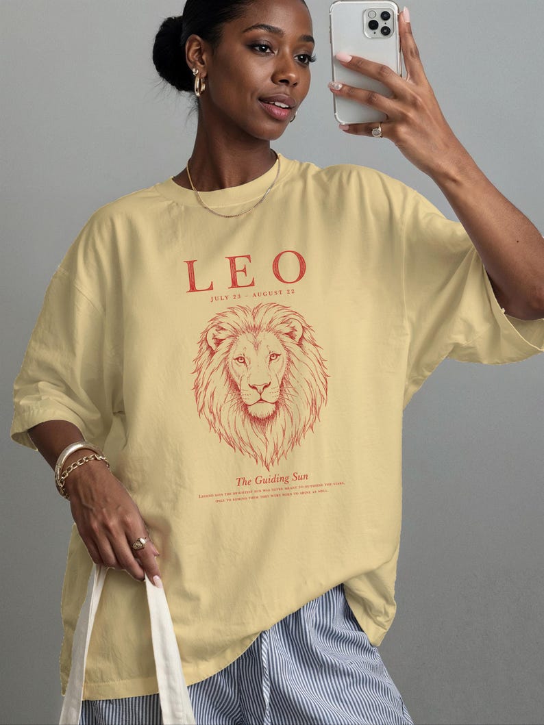 K&ouml;nnte beinhalten: Ein hellgelbes Kurzarm-T-Shirt mit einer roten Grafik eines L&ouml;wenkopfes. Der Text lautet LEO, JULY 23 - AUGUST 22 und The Guiding Sun, mit kleinerem Text darunter. Das T-Shirt hat einen Rundhalsausschnitt und eine lockere, entspannte Passform.