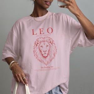 K&ouml;nnte beinhalten: Ein hellrosa T-Shirt mit einer roten Grafik eines L&ouml;wenkopfes. Der Text lautet LEO, JULY 23 - AUGUST 22 und The Guiding Sun. Das T-Shirt hat einen Rundhalsausschnitt, kurze &Auml;rmel, eine lockere Passform und ein aufgedrucktes Design auf der Vorderseite.