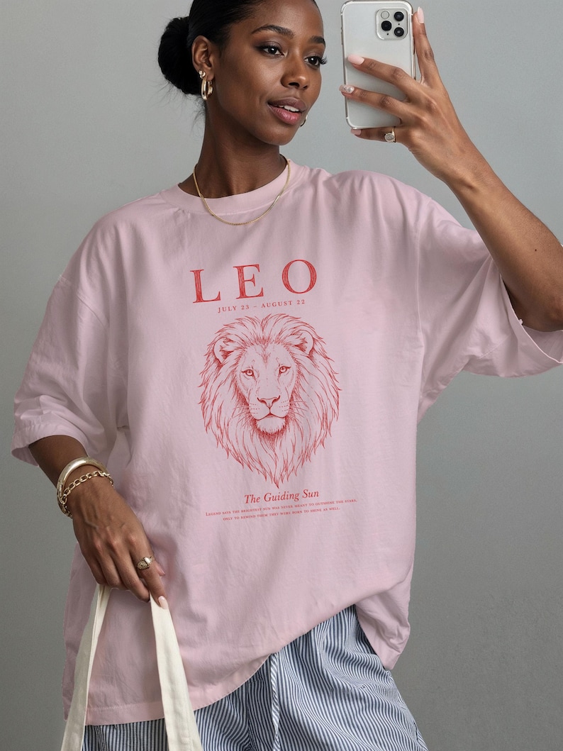 K&ouml;nnte beinhalten: Ein hellrosa T-Shirt mit einer roten Grafik eines L&ouml;wenkopfes. Der Text lautet LEO, JULY 23 - AUGUST 22 und The Guiding Sun. Das T-Shirt hat einen Rundhalsausschnitt, kurze &Auml;rmel, eine lockere Passform und ein aufgedrucktes Design auf der Vorderseite.