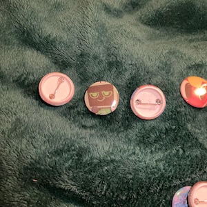 Puede incluir: Cuatro botones redondos con varios diseños. Uno tiene un fondo blanco con una ilustración de cuchara. Otro presenta un personaje con cara verde. El tercero tiene un alfiler. El último tiene un diseño colorido con rojo, amarillo y naranja.