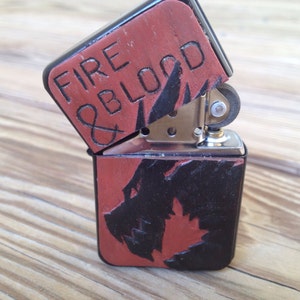 Puede incluir: Un encendedor negro y rojo con un dise&ntilde;o de drag&oacute;n y las palabras "Fire & Blood" grabadas en la parte superior.