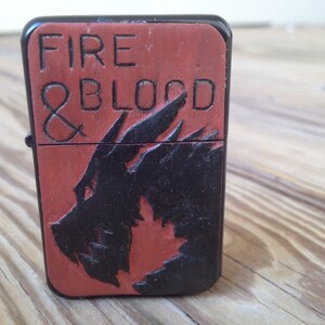 Puede incluir: Un encendedor negro y rojo con las palabras "FIRE & BLOOD" y un dise&ntilde;o de drag&oacute;n.