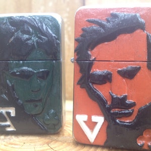 Peut inclure: Deux briquets en métal de style vintage avec des portraits de hommes sculptés. Le briquet de gauche a un fond vert et la lettre "A" en bas. Le briquet de droite a un fond orange et la lettre "V" en bas.