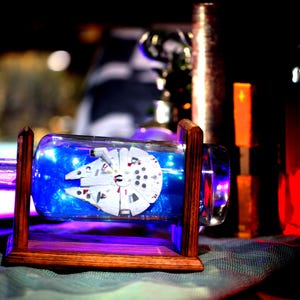 May include: A glass bottle with a wooden stand, containing a miniature model of the Millennium Falcon spaceship. The bottle is illuminated from within, creating a blue glow.