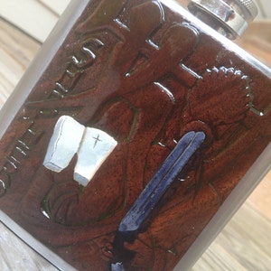 May include: A silver flask with a brown leather-like design. The design features a cross, a book, and a gun. The flask is engraved with the words "Rule the Herd".