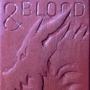 Puede incluir: Una placa de madera con las palabras "FIRE & BLOOD" talladas en ella. La placa es de color marr&oacute;n rojizo y presenta un dise&ntilde;o de drag&oacute;n tallado.