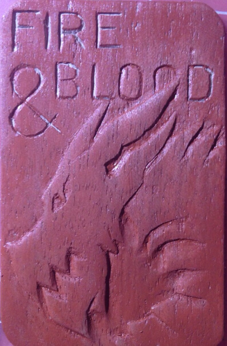 Puede incluir: Una placa de madera con las palabras "FIRE & BLOOD" talladas en ella. La placa es de color marr&oacute;n rojizo y presenta un dise&ntilde;o de drag&oacute;n tallado.