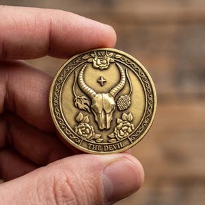 May include: A round, antique gold-colored coin featuring a bull skull with horns, roses, and the text "THE DEVIL". The coin has a decorative border and is held in a hand.