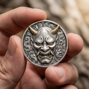 May include: A circular silver coin featuring a detailed relief of a Hannya mask with gold-colored horns, eyes, and fangs. The background of the coin is decorated with intricate floral patterns and swirling cloud motifs in a metallic silver finish.