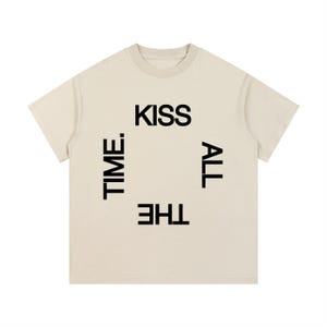 May include: Cream-colored t-shirt with the words "KISS ALL THE TIME." in black block letters. The text is arranged in a square around a central open space. The shirt has short sleeves and a crew neck.