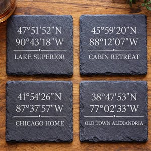 May include: Four slate coasters with white text and coordinates. The coasters read "Lake Superior", "Cabin Retreat", "Chicago Home", and "Old Town Alexandria". A glass of amber liquid sits in the upper left corner.