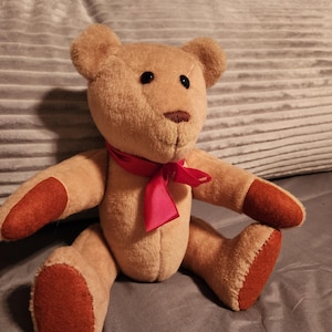 May include: A plush teddy bear with tan fur, dark brown eyes, and a brown nose. The bear has red-brown paws and a red ribbon tied around its neck. The bear is sitting on a gray surface.