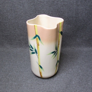 Puede incluir: Un jarrón de cerámica blanco con una parte superior ondulada única. El jarrón presenta un diseño de bambú con tallos amarillos, hojas verdes y un degradado rosa en la parte superior. El jarrón mide unos 18 cm de alto.