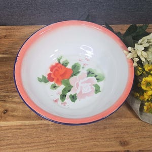 Puede incluir: Un cuenco de esmalte blanco con borde rosa y ribete azul. El cuenco presenta un diseño floral con rosas rojas y rosas y hojas verdes. El cuenco está colocado sobre una superficie de madera.