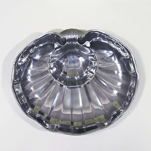 Puede incluir: Una fuente plateada con forma de concha. El plato tiene una sección exterior grande y festoneada y un cuenco central más pequeño. La superficie refleja la luz, resaltando el diseño detallado.