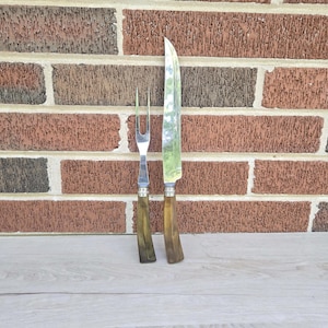 Può includere: Un set di coltello e forchetta da intaglio con manici marroni e lame e rebbi argentati. Il coltello è lungo e sottile, mentre la forchetta ha due rebbi. Il set è esposto contro un muro di mattoni.