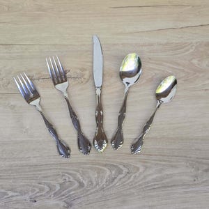 Puede incluir: Un juego de cinco utensilios plateados: dos tenedores, un cuchillo y dos cucharas. Los utensilios tienen mangos ornamentados y están colocados sobre una superficie de madera clara. La cubertería es para comer.