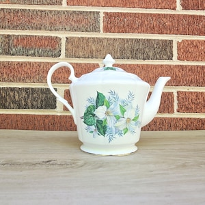 May include: White porcelain teapot with a lid, handle, and spout. The teapot is decorated with a floral design featuring white flowers and green leaves. The teapot sits on a wooden surface in front of a brick wall.