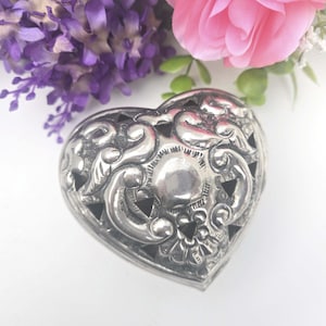 May include: A silver heart-shaped trinket box with ornate floral and scrollwork detailing. The box has a hinged lid and is surrounded by purple and pink flowers, creating a romantic and vintage aesthetic.