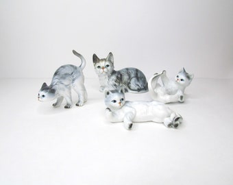 May include: Four ceramic cat figurines in various poses. The cats are primarily white and gray, with blue eyes and pink noses. One cat is arched, another is lying down, and two are in playful positions.