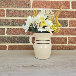 May include: A cream-colored ceramic pitcher with two black stripes, filled with white and yellow artificial flowers. The pitcher has a handle and is set against a brick wall and a wooden surface.