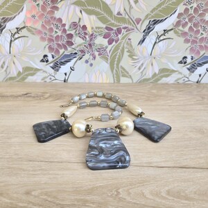May include: A vintage necklace featuring a strand of gray and cream-colored beads with gold accents. The necklace has large, marbled gray and black pendant shapes and large cream-colored faux pearls.