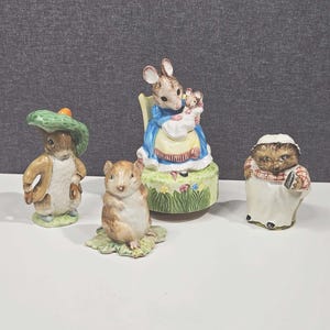 Puede incluir: Cuatro figuras de cerámica de personajes de cuentos. Un conejo con un sombrero verde, un pequeño hámster marrón, un ratón sosteniendo un bebé ratón y un erizo con un delantal blanco y una bufanda a cuadros rojos y blancos.