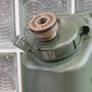 May include: Close-up of an olive green water container with a tan cap. The cap is labeled "SUBLIME FABWORKS" in black text. The container has a hinged lid and a black rubber seal.