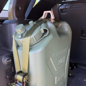 May include: An olive green Scepter 20L water jerry can secured with a green nylon ratchet strap. The plastic container features a screw-top lid, a molded handle, and embossed text reading Scepter, 20L, and Water on the front side surface.