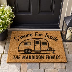 May include: A rectangular tan coir doormat featuring a black graphic of a vintage camper trailer. The text S'more Fun Inside is printed above the camper, and the text THE MADDISON FAMILY is printed below the camper in a bold black font.