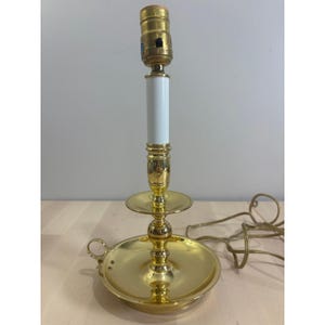 May include: A polished brass candlestick lamp featuring a wide circular base with a small side handle, a central stem with a white cylindrical sleeve, and a brass light socket at the top, accompanied by a thin electrical cord with a plug.