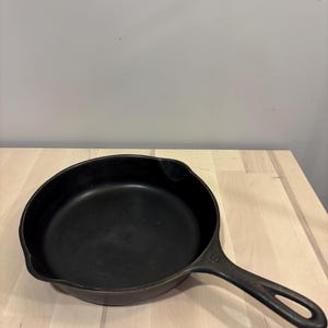 May include: A black cast iron skillet with a round cooking surface, two side pouring spouts, and a long handle featuring a hole at the end for hanging. The number 6 is embossed on the top of the handle near the base of the pan.