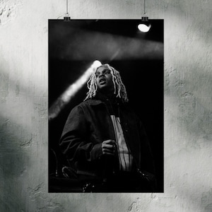 May include: Black and white poster print of a person with long, light-colored dreadlocks, wearing a dark jacket and a striped shirt. The person is looking upwards, with a spotlight shining down. The poster is hanging on a textured wall.