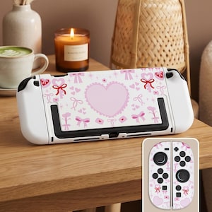 May include: A white Nintendo Switch console with a custom skin featuring a pink heart, bows, and floral patterns. The design is repeated on the attached controller. A candle, coffee cup, and mouse are also visible.