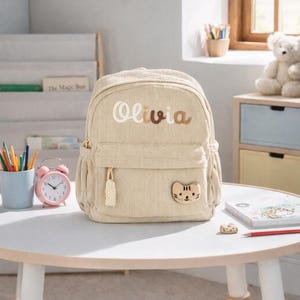 Könnte beinhalten: Ein beigefarbener Rucksack aus Cord mit dem Namen Olivia, der in braunen Buchstaben auf der Vorderseite eingestickt ist. Die Tasche verfügt über eine Reißverschlusstasche vorne mit einem kleinen, hellen Katzenaufnäher unten rechts.