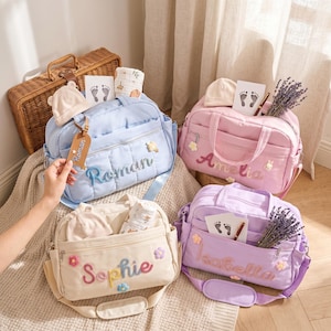 May include: Four quilted diaper bags in light blue, pink, cream, and lavender, each featuring embroidered names: Roman, Amelia, Sophie, and Isabella. The bags include small floral accents, baby hats, footprint cards, and lavender sprigs.