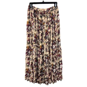 Peut inclure: Longue jupe crème à motif floral dans les tons de violet, bordeaux et jaune. La jupe est plissée et suspendue à un cintre noir. Le tissu semble léger et fluide, adapté aux températures plus chaudes.