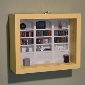 Puede incluir: Diorama de estantería en miniatura con marco amarillo. El interior presenta estantes blancos llenos de libros diminutos, un pequeño televisor y objetos decorativos. También se ven un CD, un taburete pequeño y un cubo de basura.