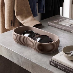 May include: A taupe ribbed sunglasses holder containing sunglasses, placed on a concrete surface. Books with the text "HIGH DESIGN" are nearby. A tan coat and denim jacket hang in the background. The holder is likely made of plastic or a similar material.